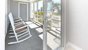 Un balcon avec des fauteuils à bascule blancs et une porte vitrée donnant sur une vue de palmiers.