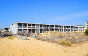 Un edificio frente a la playa con techo plano, varios balcones y un paseo de madera que lleva a la entrada.