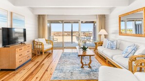 A spacious living room with a wooden floor, a television, a sofa, a coffee table, and a view of the beach through large glass doors. - Beach Place 14 2 Bedroom Townhouse (Ocean City)