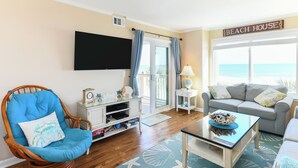 Una sala de estar con una silla de mimbre, un sofá, una mesita de centro y un televisor montado en la pared.