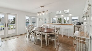 A dining area with a wooden table and white chairs, a kitchen with white cabinets, and a balcony with a view of houses.