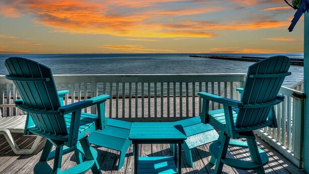 Eine Holzterrasse mit zwei türkisfarbenen Adirondack-Stühlen und einem kleinen Tisch, mit Blick auf ein Gewässer unter einem Sonnenuntergangshimmel.
