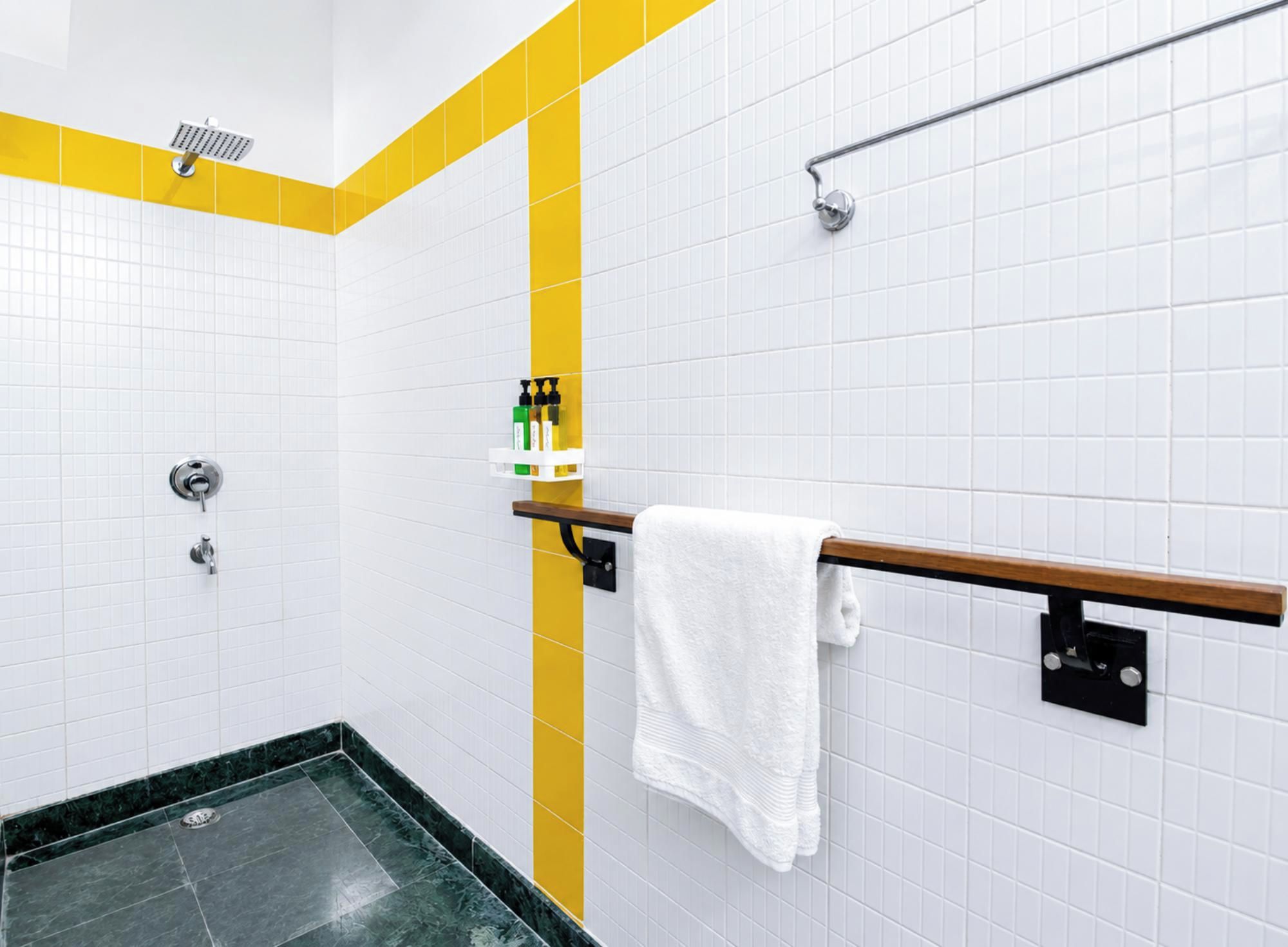 Une salle de bain avec des carreaux blancs, un mur d’accent jaune, une douche avec un pommeau de douche amovible, un porte-serviettes avec une serviette blanche et une étagère avec des articles de toilette.