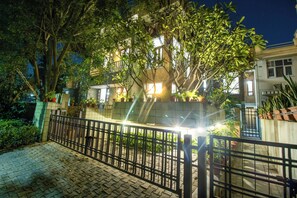 Un área residencial cerrada con un camino pavimentado, árboles y un edificio con ventanas iluminadas por la noche.