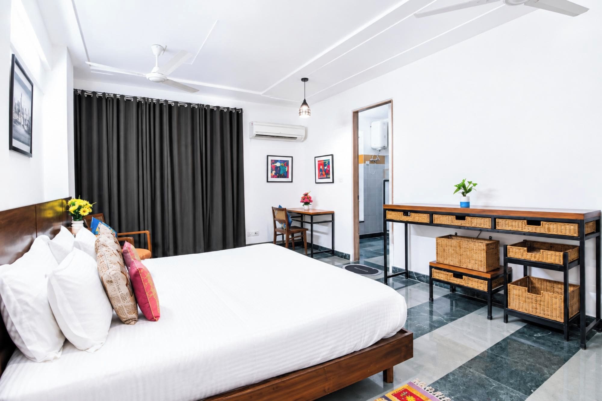 A hotel room with a bed, a desk, a chair, a small table, a potted plant, and artwork on the walls.