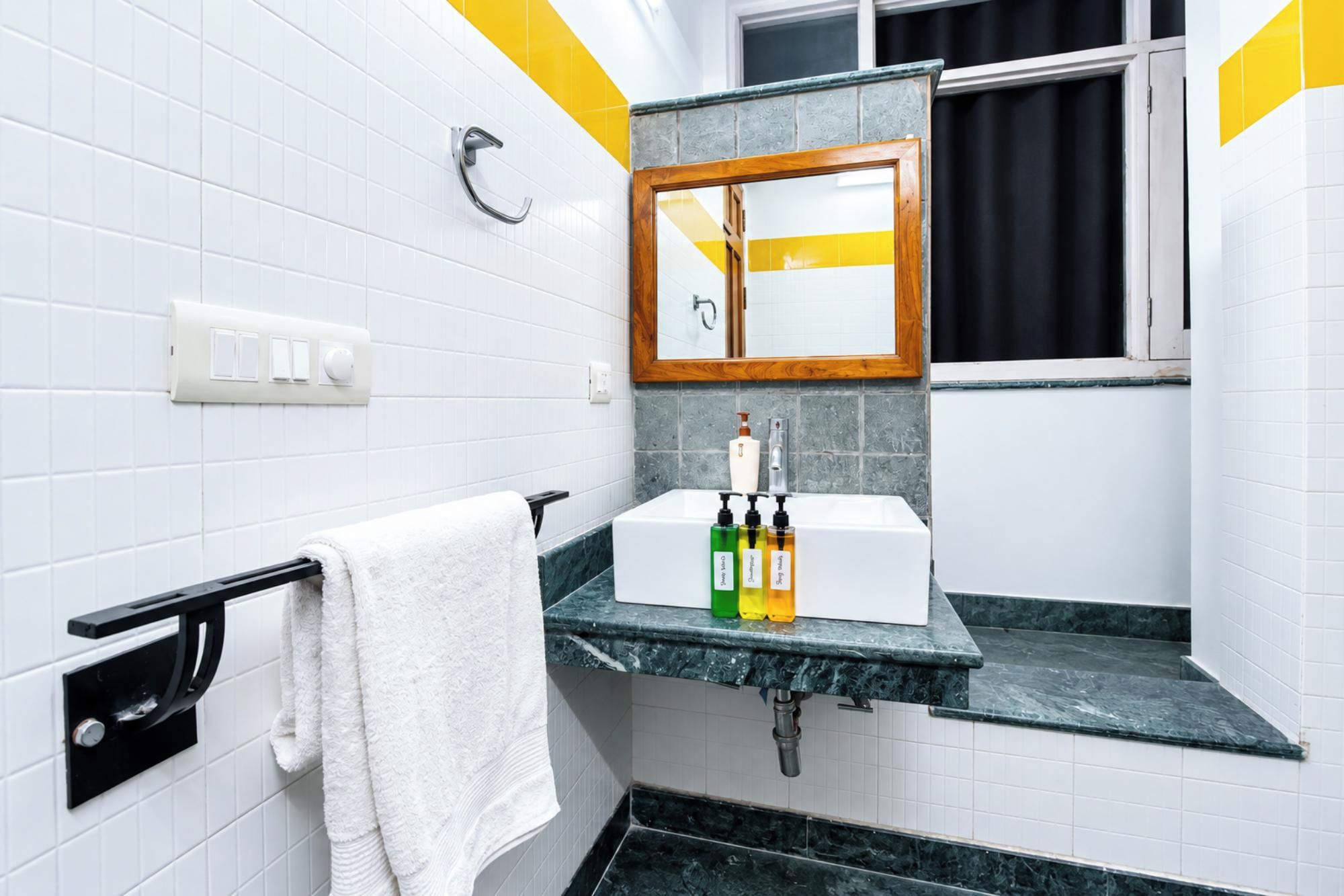 A bathroom with white tiled walls, a black towel rack, a mirror with a wooden frame, and a sink with soap dispensers.