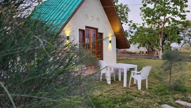 A small house with a green roof, a wooden door, and a white table with two chairs in the yard.