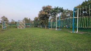 A grassy field with a playground featuring a climbing net, a colorful fence, and trees in the background.