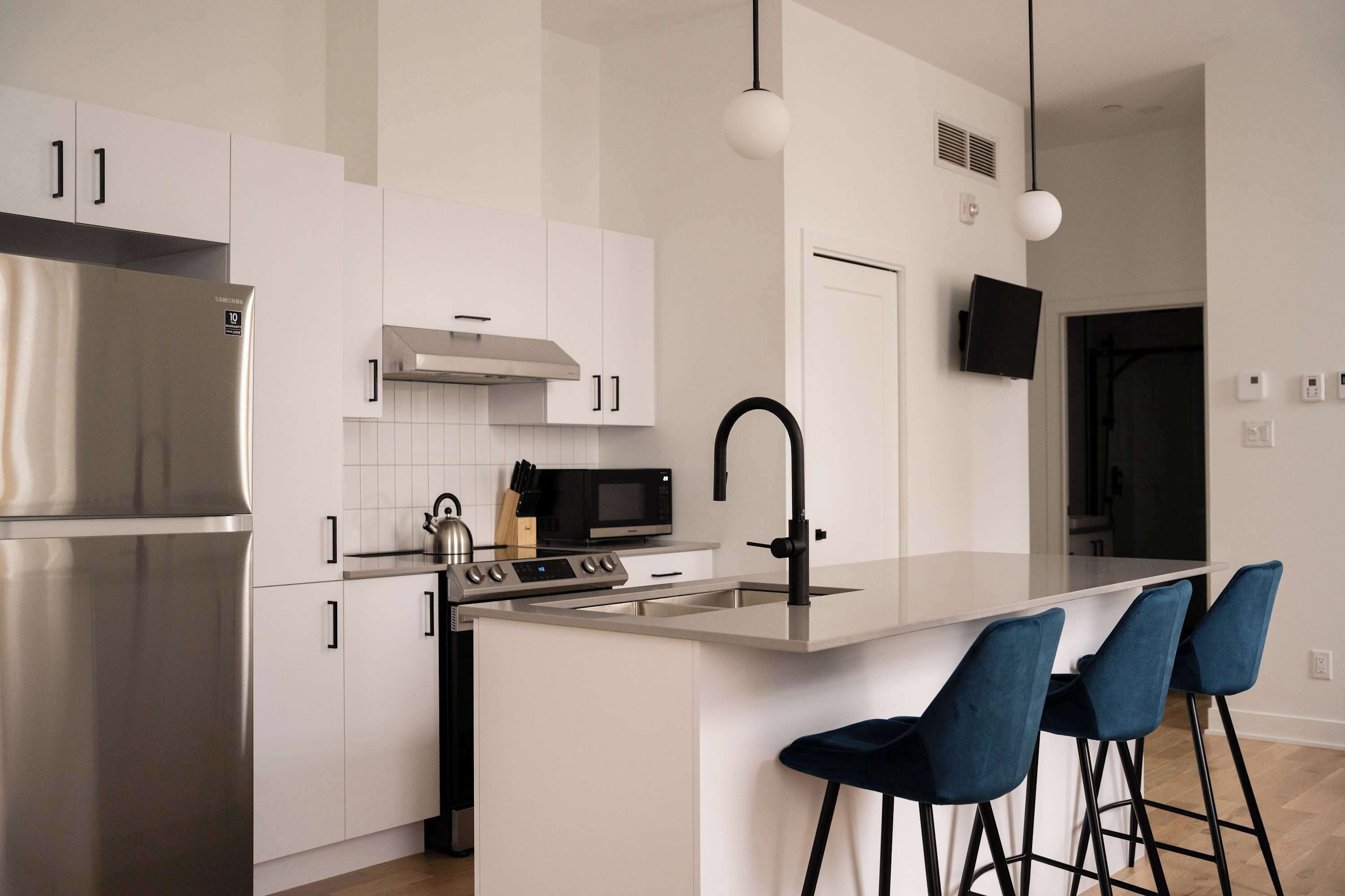 Een moderne keuken met witte kastjes, roestvrijstalen apparatuur, een centraal eiland met barkrukken en hanglampen.