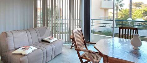 A modern living room with a sofa, wooden chairs, a glass table, and a large sliding glass door leading to a balcony with a view of palm trees.