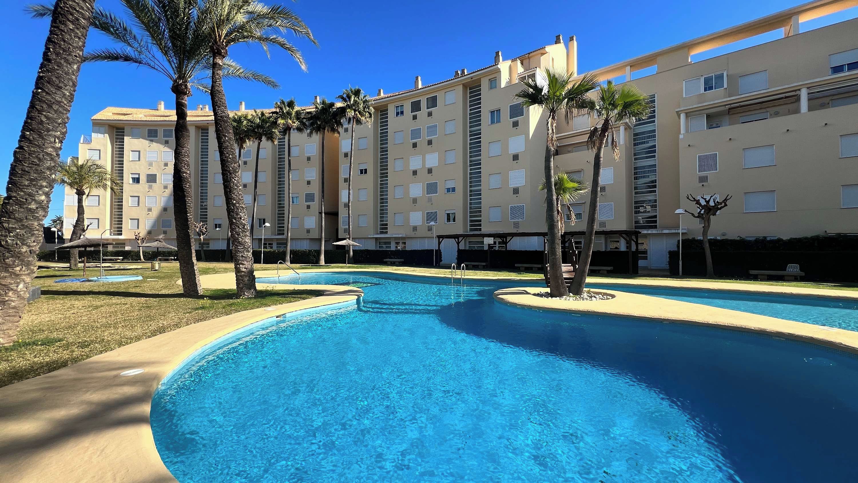 A swimming pool with a curved design, surrounded by palm trees and a multi-story building with balconies.