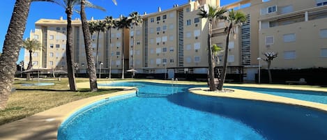 A swimming pool with a curved design, surrounded by palm trees and a multi-story building with balconies.