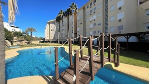 A swimming pool with wooden deck and rope barriers, surrounded by a multi-story building with balconies and palm trees.