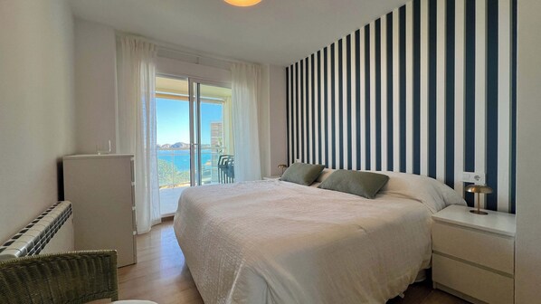 A bedroom with a large bed, a nightstand, a lamp, and a view of the sea through a sliding door.