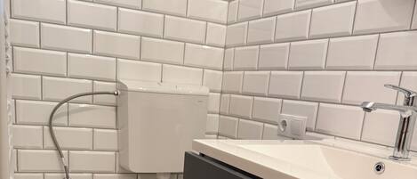 Un baño con inodoro, lavamanos y ducha. Las paredes están revestidas de baldosas blancas y el suelo tiene un diseño con baldosas.