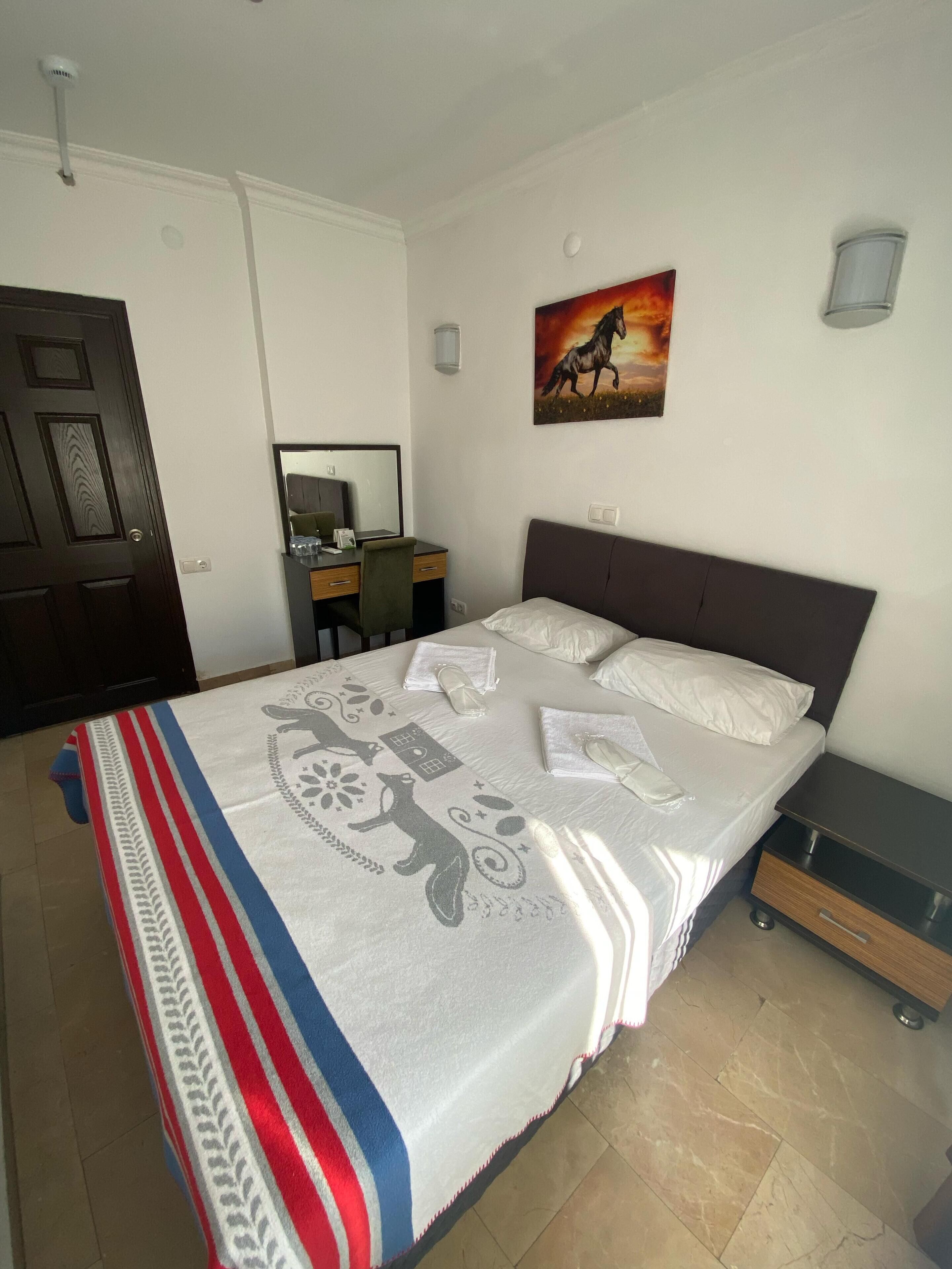 A neatly made bed with a patterned blanket, a desk with a chair, a painting on the wall, and a door.