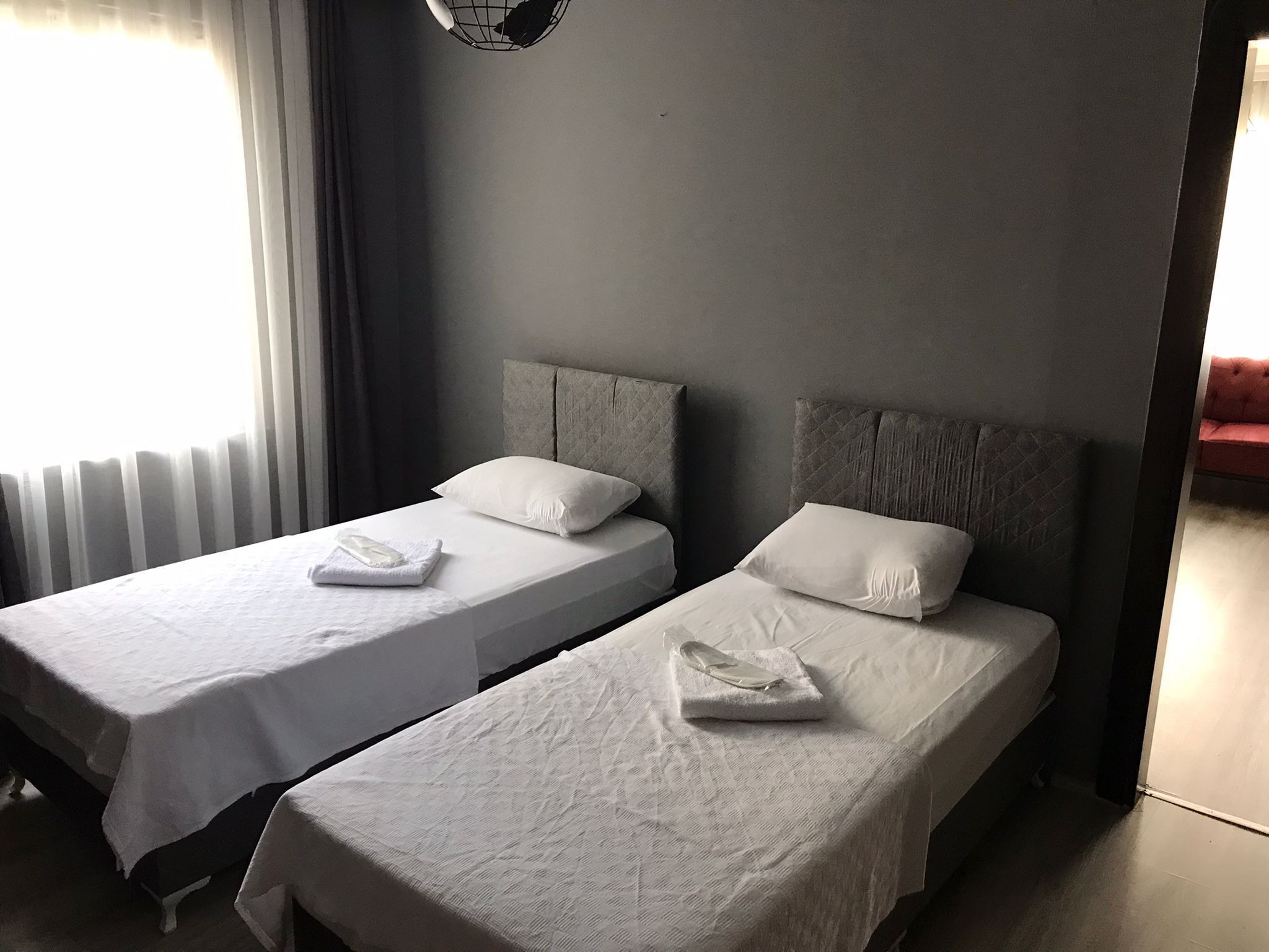A hotel room with two single beds, each with a white bedspread and pillow, a grey headboard, and a window with sheer curtains.