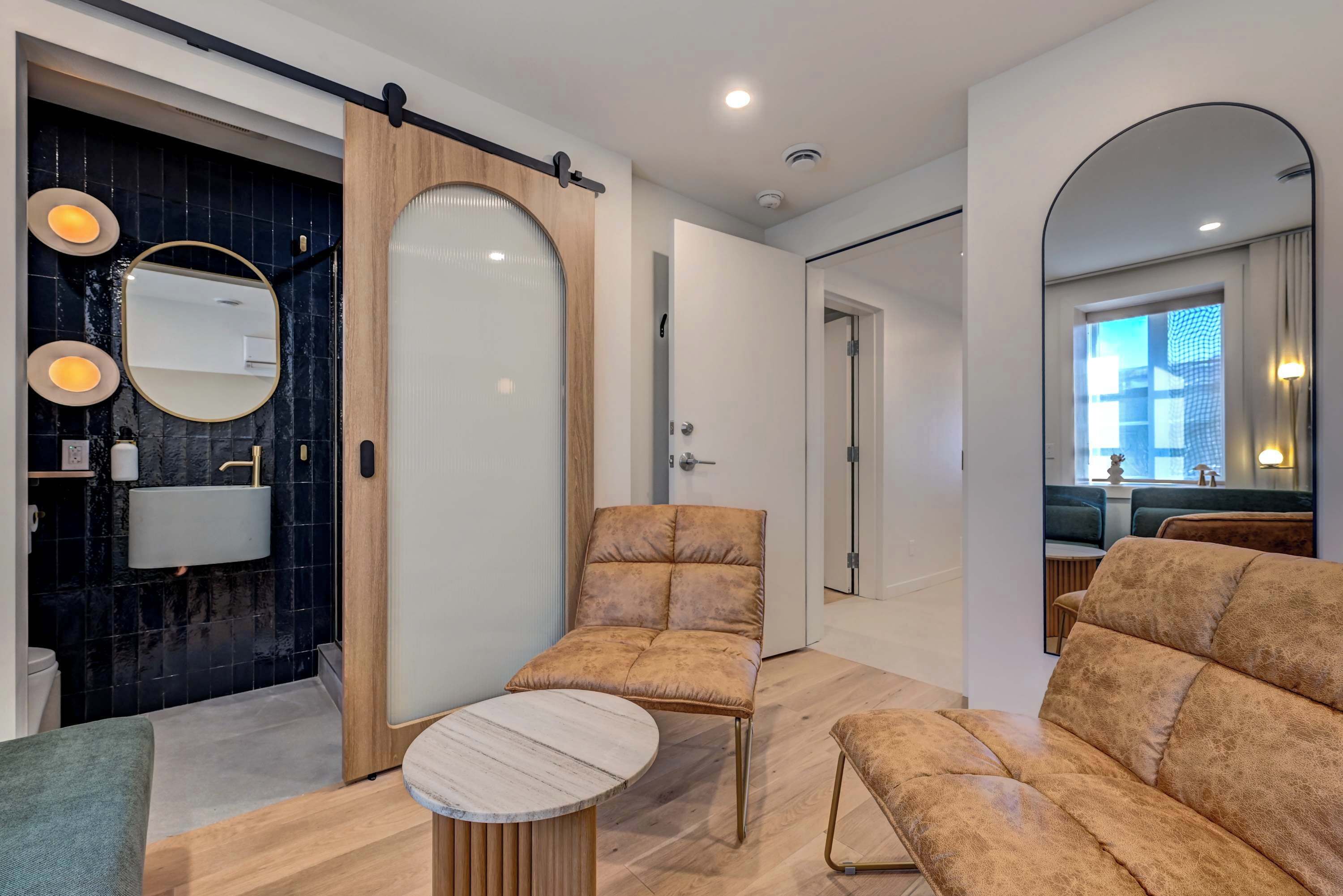Une salle de bain moderne avec un mur en marbre noir, un miroir rond et une porte en bois. Le salon est aménagé avec deux canapés beige et une petite table ronde.