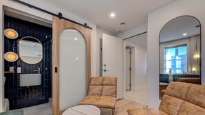Un baño moderno con pared de mármol negro, un espejo redondo y una puerta de madera. En la zona de estar hay dos sofás color beige y una pequeña mesa redonda.