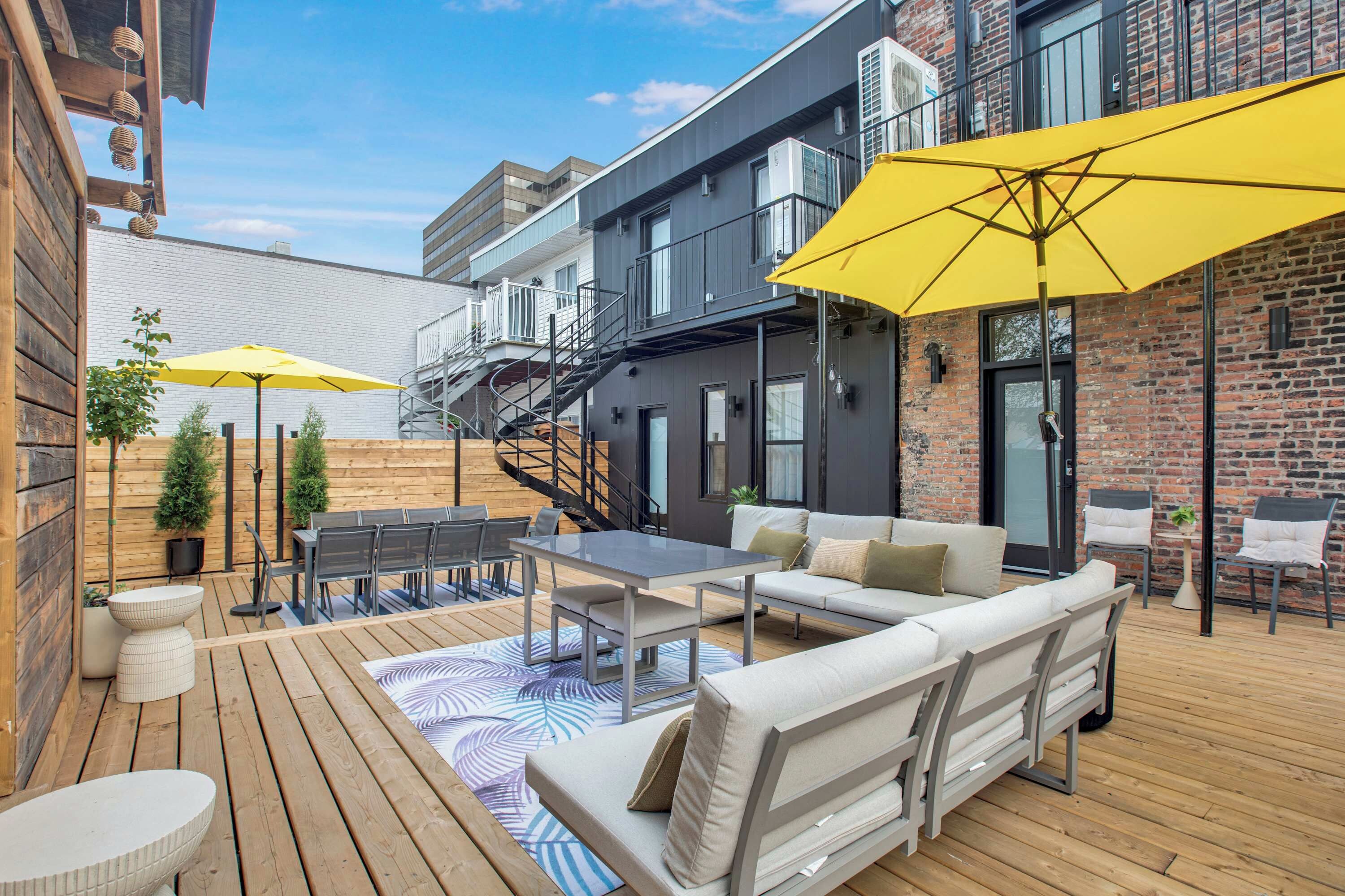 Une terrasse extérieure avec une plateforme en bois, une table à manger, des chaises et un canapé. Deux parasols jaunes offrent de l’ombre.