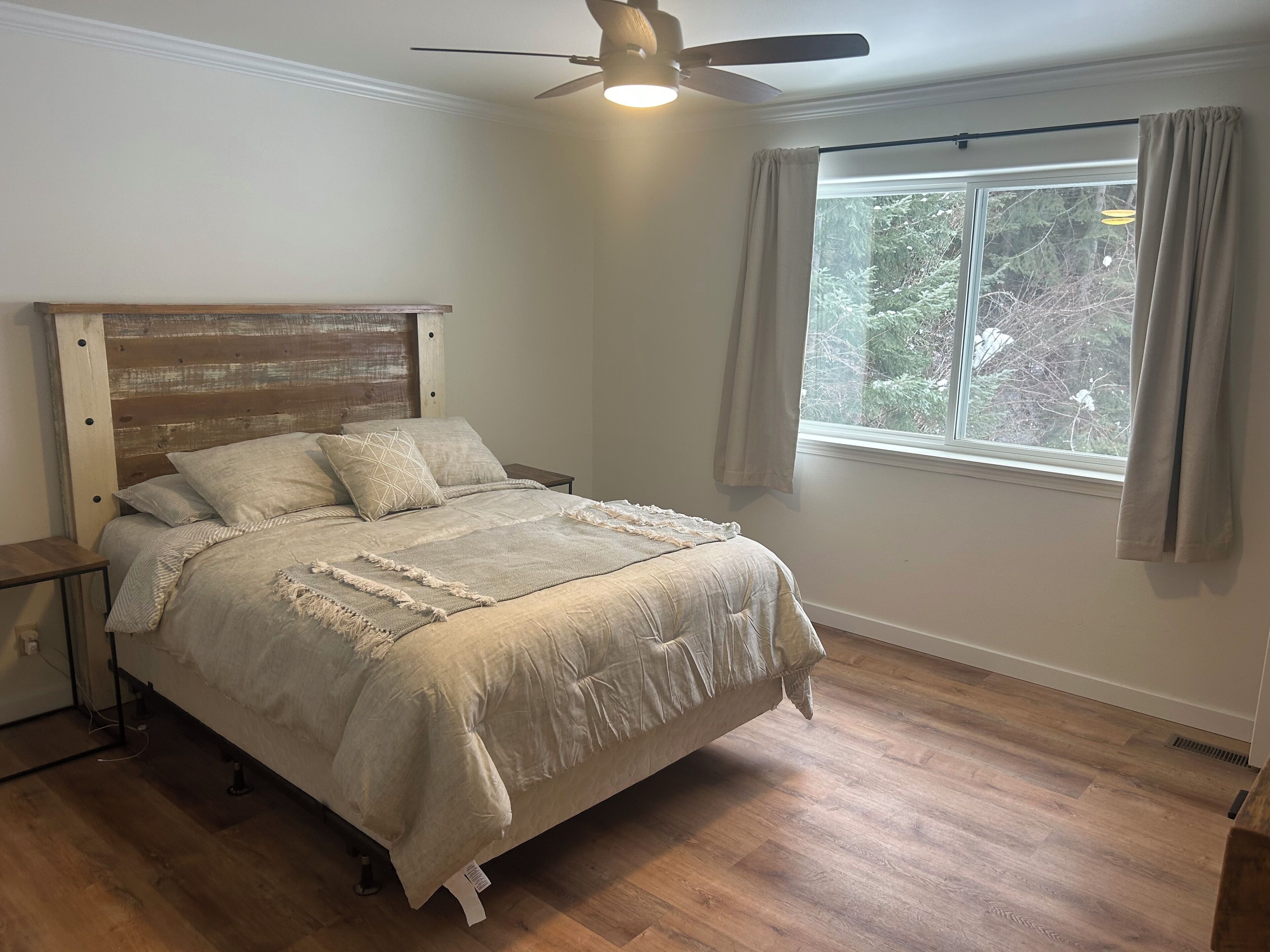 Ein Schlafzimmer mit einem hölzernen Kopfteil, einem Bett mit einer beigefarbenen Tagesdecke, einem Nachttisch, einem Fenster mit Vorhängen und einem Deckenventilator.
