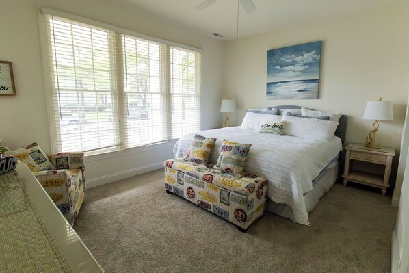 A bedroom with a bed, two bedside lamps, a painting of a beach, and a bench with beach-themed cushions.