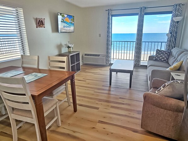 A living room with a wooden floor, a dining table with chairs, a sofa, a TV mounted on the wall, and a view of the ocean through large windows. - Direct Oceanfront and Boardwalk Condo! Decatur House 510 (Ocean City)