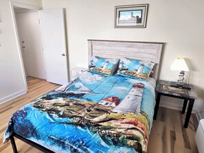 A bedroom with a bed featuring a nautical-themed bedspread, a nightstand with a lamp, and a framed picture on the wall. - Direct Oceanfront and Boardwalk Condo! Decatur House 510 (Ocean City)