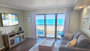 A living room with a large TV, a sofa, a coffee table, and a view of the beach through the sliding glass door.