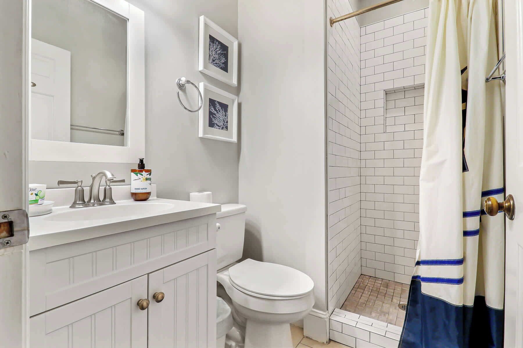 Un baño con tocador blanco, inodoro, ducha con azulejos blancos y un cuadro en la pared.
