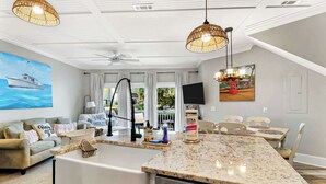 A modern kitchen with a central island, a dining area with chairs, a ceiling fan, and a large window with curtains. - The Beach is Calling-this 3bd Villa is the answer! (Hilton Head Island)