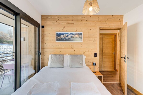 Un dormitorio con panel de madera en la pared, cama con ropa de cama blanca, mesita de noche, lámpara y un cuadro de montañas.