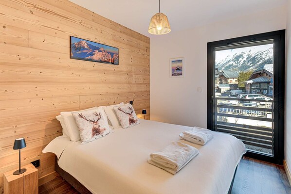 Un dormitorio con cabecera de madera, cama con ropa de cama blanca, lámpara y vista a la montaña a través de un ventanal.