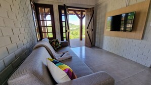 A modern living room with a grey sofa, colorful cushions, a mounted TV, and a view of greenery through open doors. - CalaRosa . Condomínio (Imbituba)