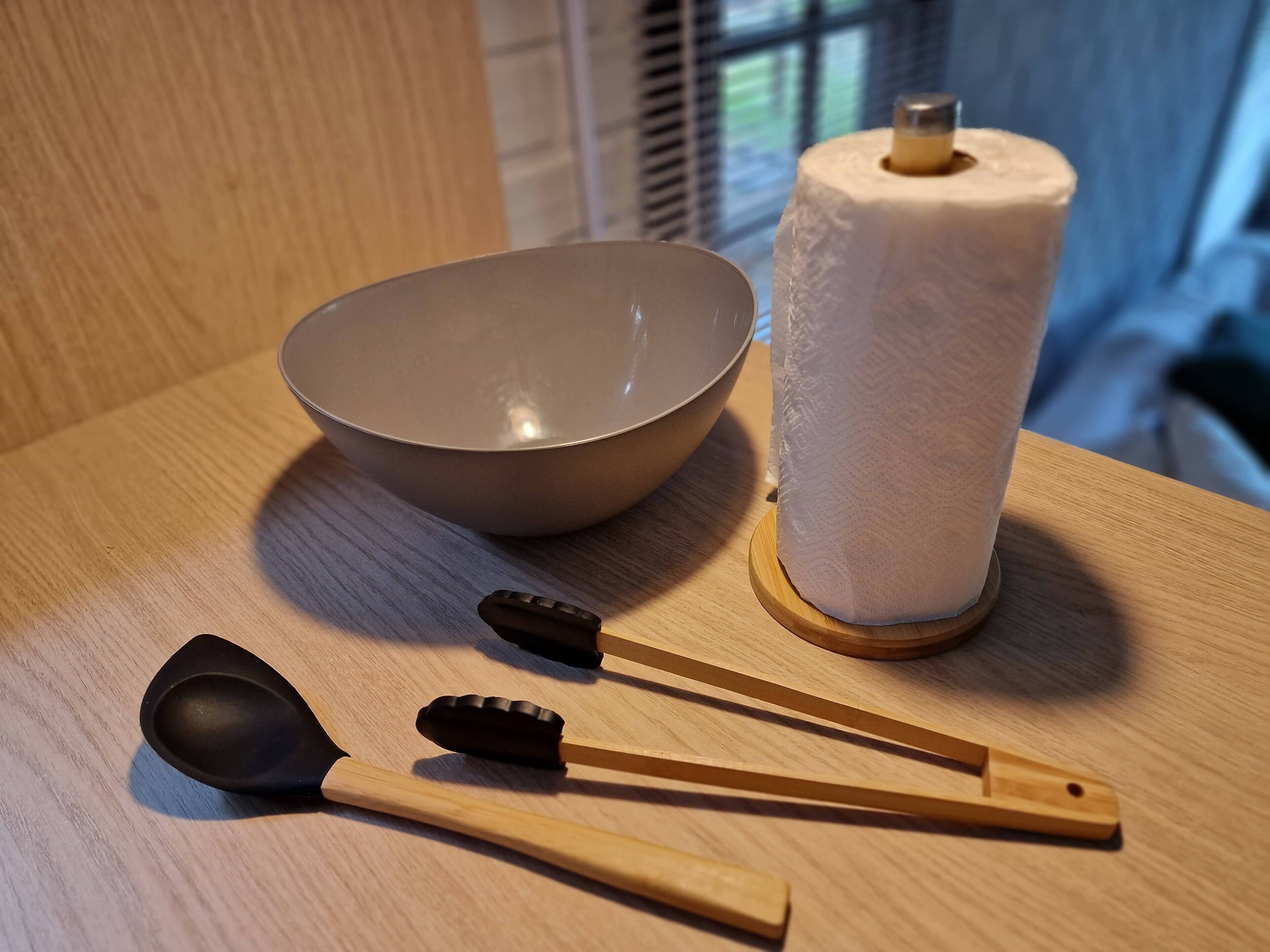 A wooden table with a bowl, a roll of paper towels, and two wooden utensils.