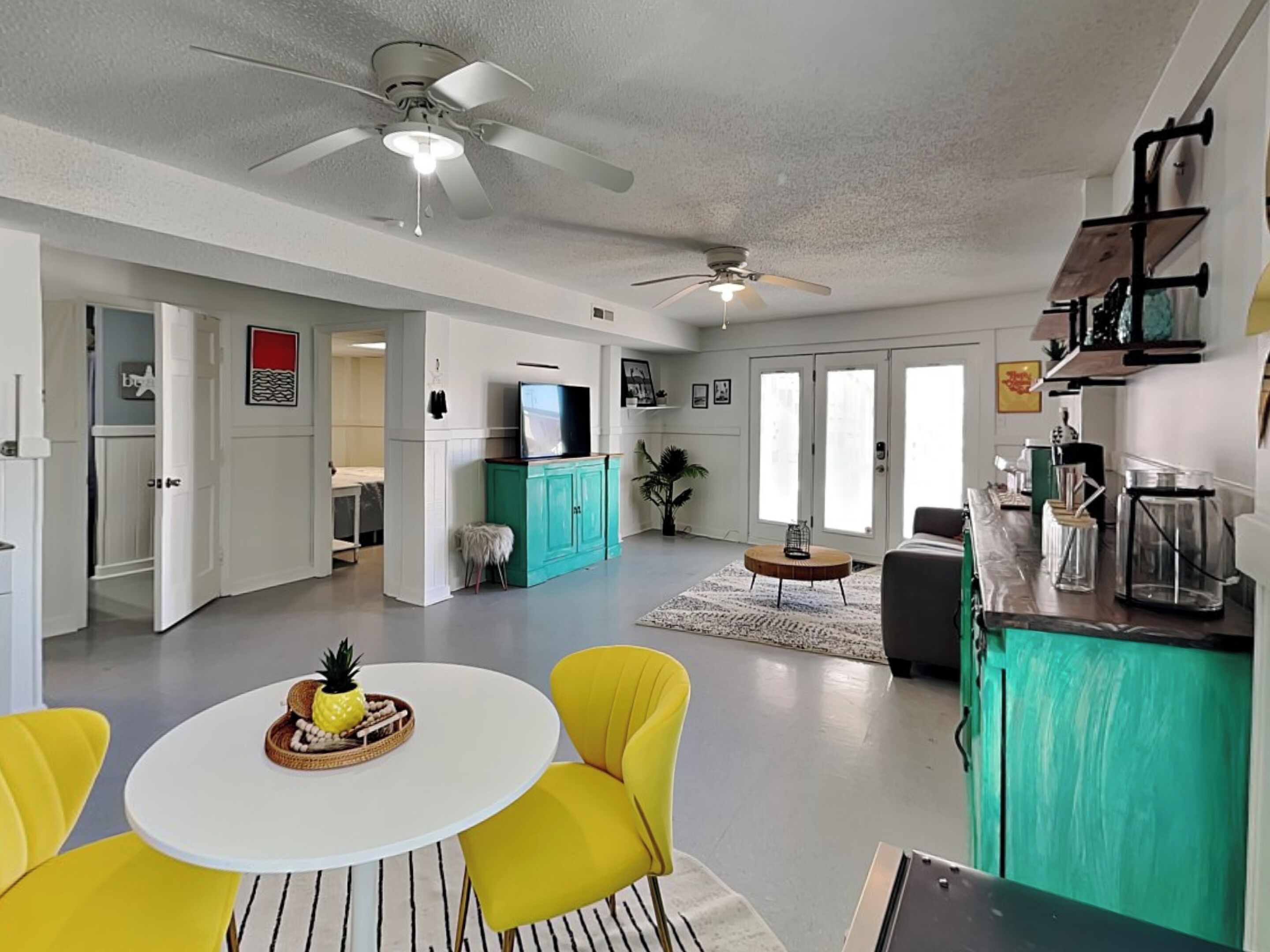 A spacious living area with a dining table, yellow chairs, a teal cabinet, and a ceiling fan.