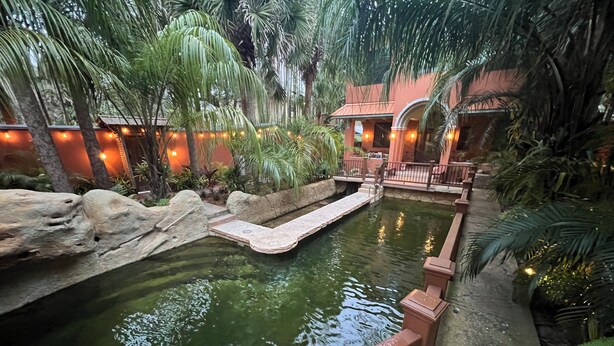 Un espace intérieur paisible avec un élément aquatique, agrémenté d’un pont en pierre, entouré d’une végétation luxuriante et éclairé d’une lumière douce et chaleureuse.