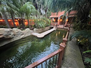 Una tranquila fuente interior con un puente de piedra, rodeada de exuberante vegetación e iluminación ambiental cálida.