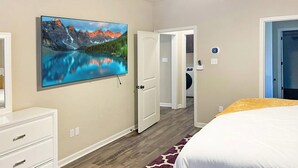 A hotel room with a large TV on the wall, a bed with a white comforter, a wooden floor, and a door leading to a laundry area. - Houma Way From Home with Arcade & EV Charger (Houma)