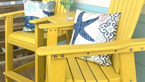 Deux chaises Adirondack jaunes, dotées de coussins à motifs, sont placées sur une terrasse en bois, près d’une maison dont le revêtement extérieur est bleu et les bordures, blanc.