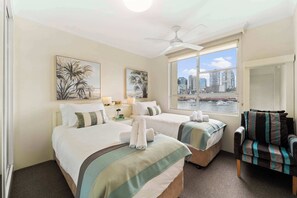 A hotel room with two beds, a chair, a window with a city view, and a ceiling fan.