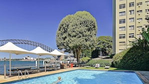 Une piscine avec une personne qui nage, un grand arbre et un bâtiment doté de nombreuses fenêtres.