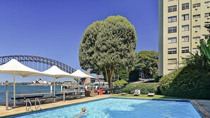A swimming pool with a person swimming, a large tree, and a building with multiple windows. - Harbourside 43 (McMahons Point)