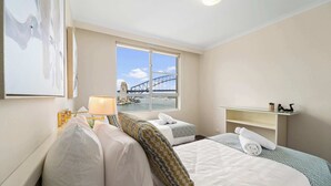 A hotel room with a bed, a bedside table with a lamp, a window with a view of a bridge, and a shelf with decorative items. - Harbourside 43 (McMahons Point)
