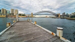 A wooden pier leading to a bridge over water with a city skyline in the background. - Harbourside 43 (McMahons Point)