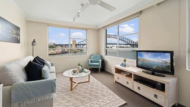 A modern living room with a sofa, a coffee table, a TV on a wooden stand, and large windows with a view of a bridge. - Harbourside 43 (McMahons Point)