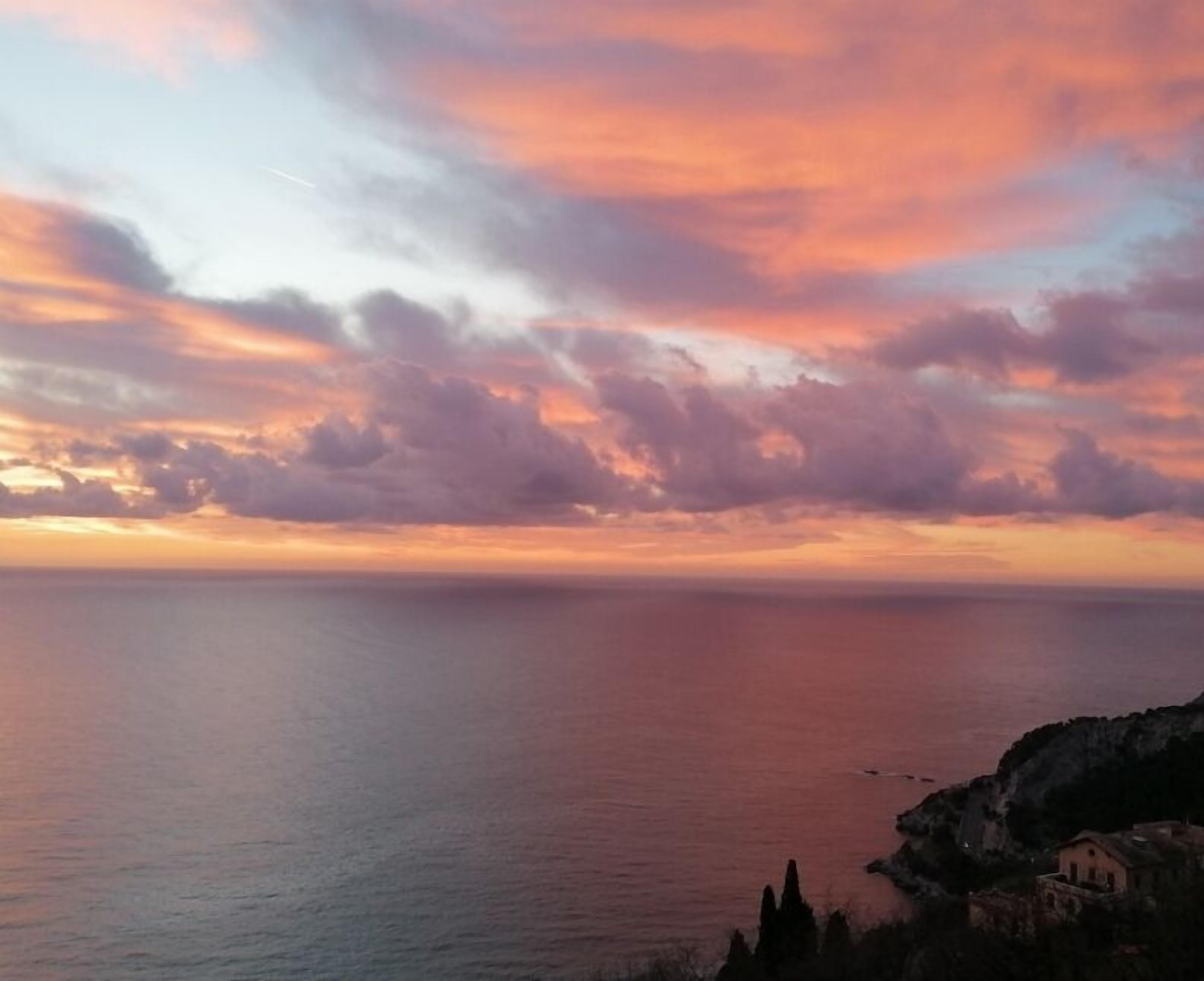 Un paisaje costero con atardecer sobre el mar, acantilados y un edificio pequeño.