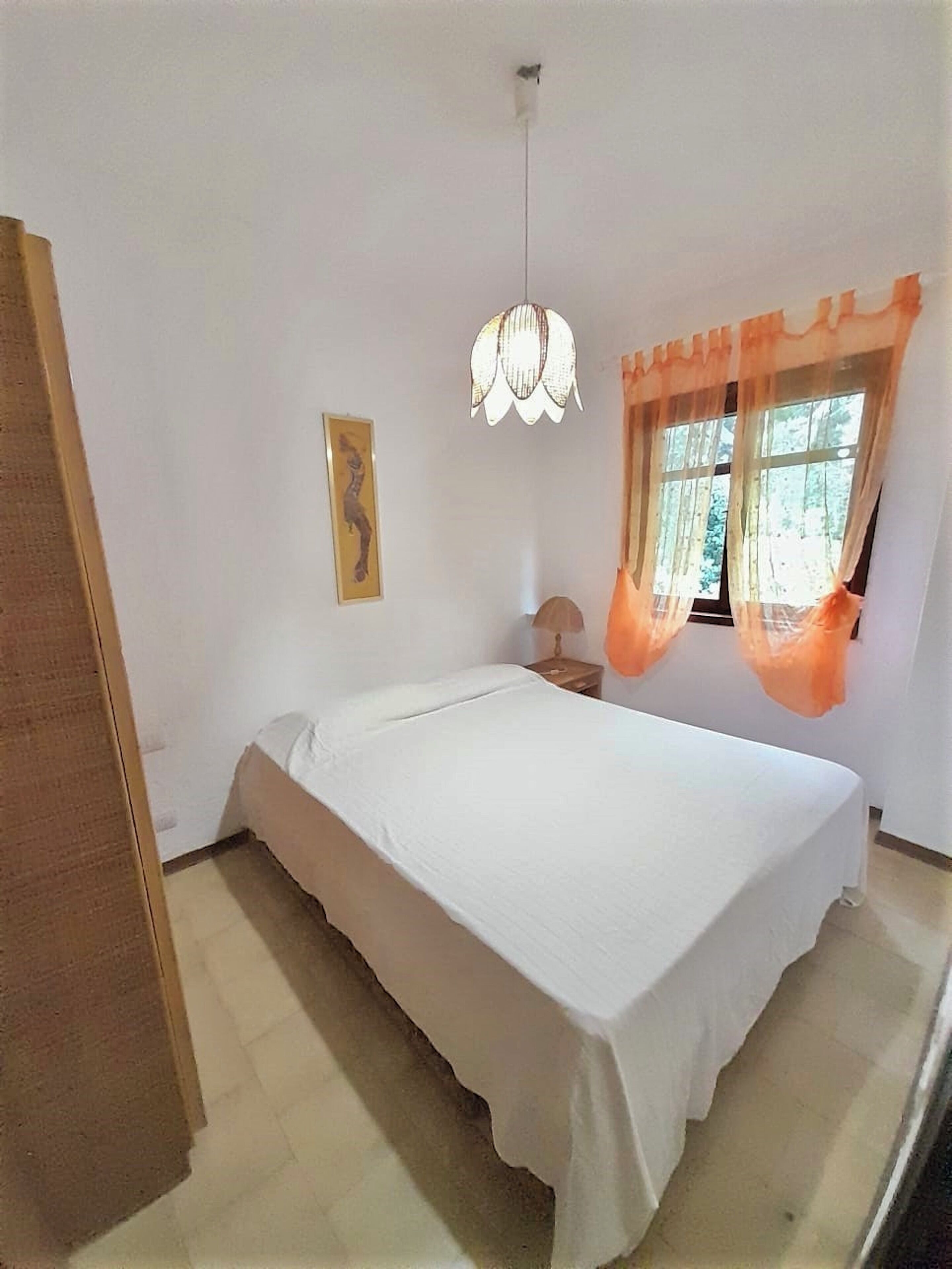 A bedroom with a single bed, a hanging light fixture, a window with curtains, and a framed picture on the wall.