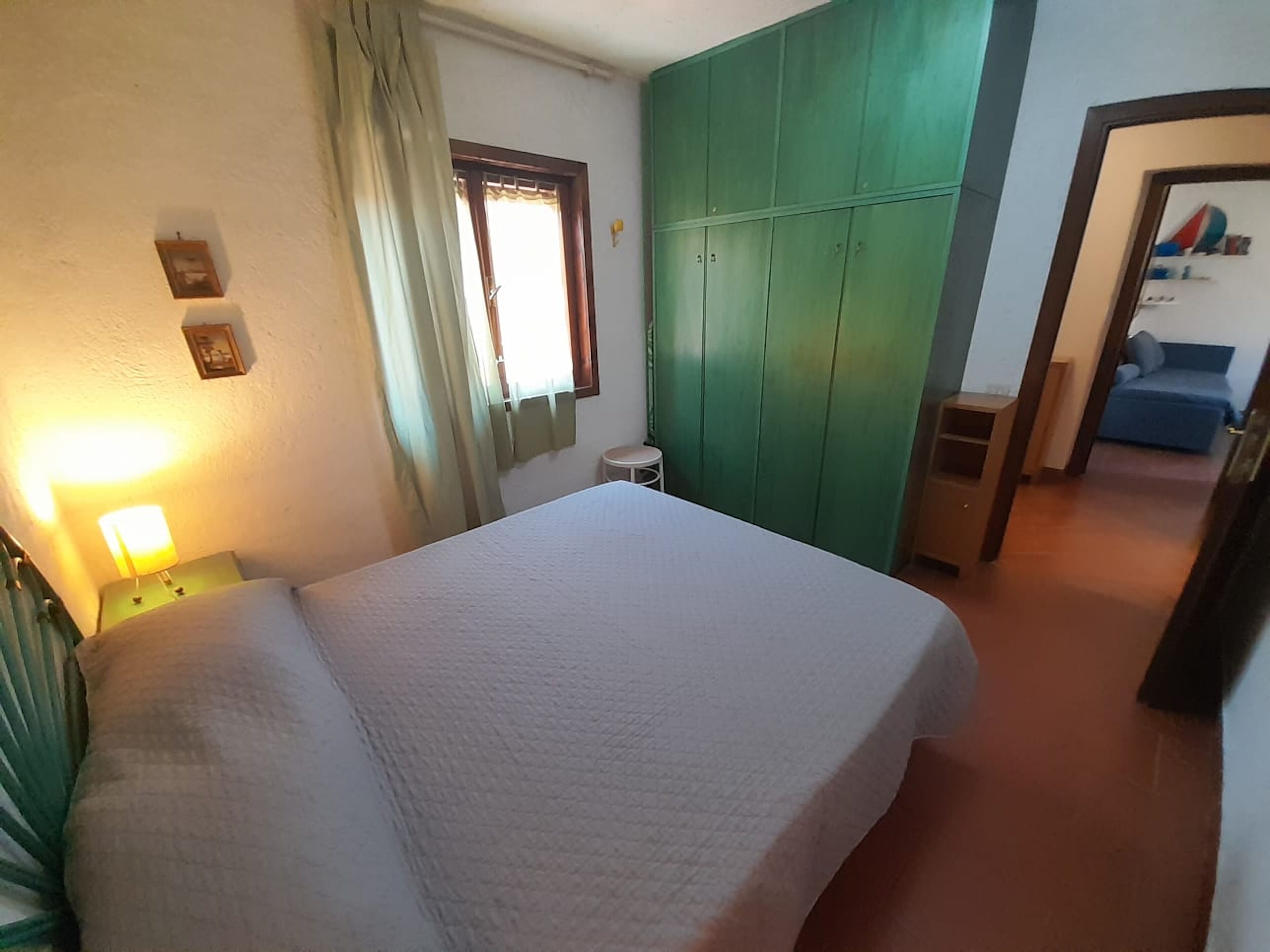 Un dormitorio con cama, una mesita de noche, un armario verde, una ventana con cortinas y una puerta que da a otra habitación.