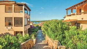 A narrow stone pathway flanked by lush greenery and traditional buildings with balconies, leading towards a clear blue sky.